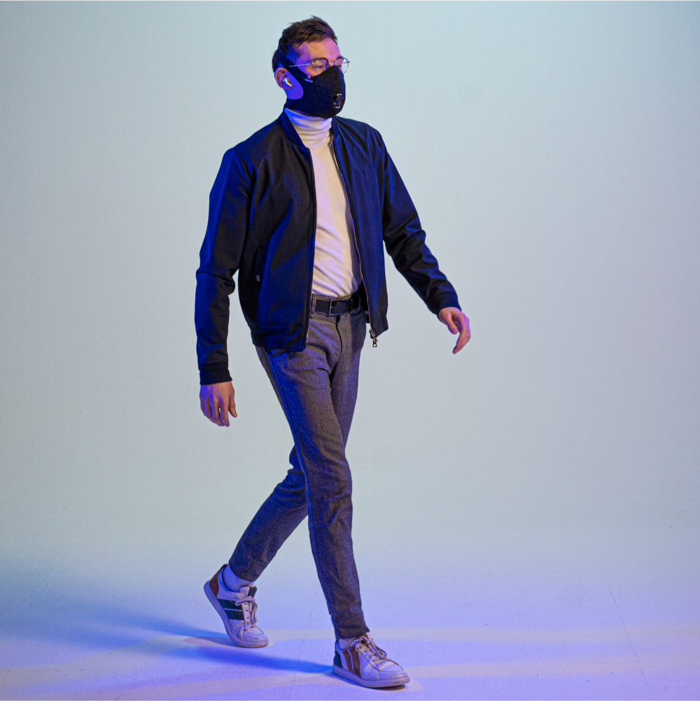 man walking while wearing a travel anti-pollution mask, a cap, and headphones, with his arms crossed and looking to the side