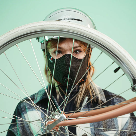 Woman in a helmet wearing a black R-PUR mask, seen through a bicycle wheel.