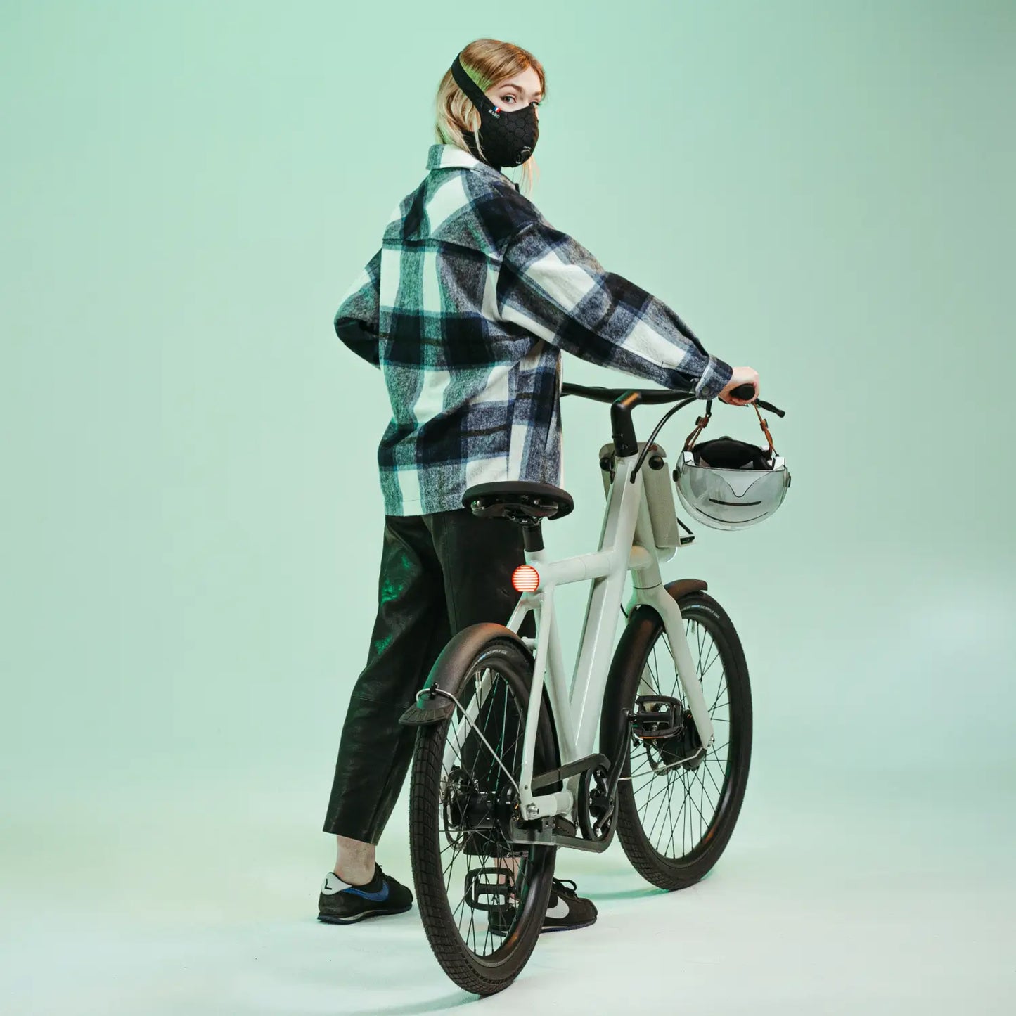 Woman in a checkered jacket wearing a black R-PUR mask, standing with a bike and holding her helmet.