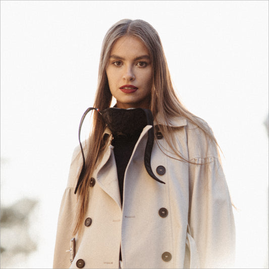 Woman facing the camera outdoors, R-PUR mask hanging at the neck over a black sweater and beige coat, ready for travel mask use