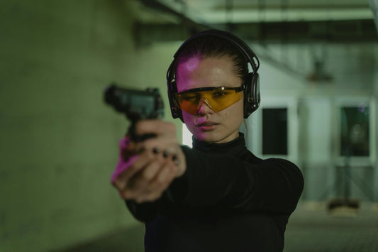 Femme portant des lunettes de protection et un casque antibruit, visant avec une arme de poing dans un stand de tir intérieur.