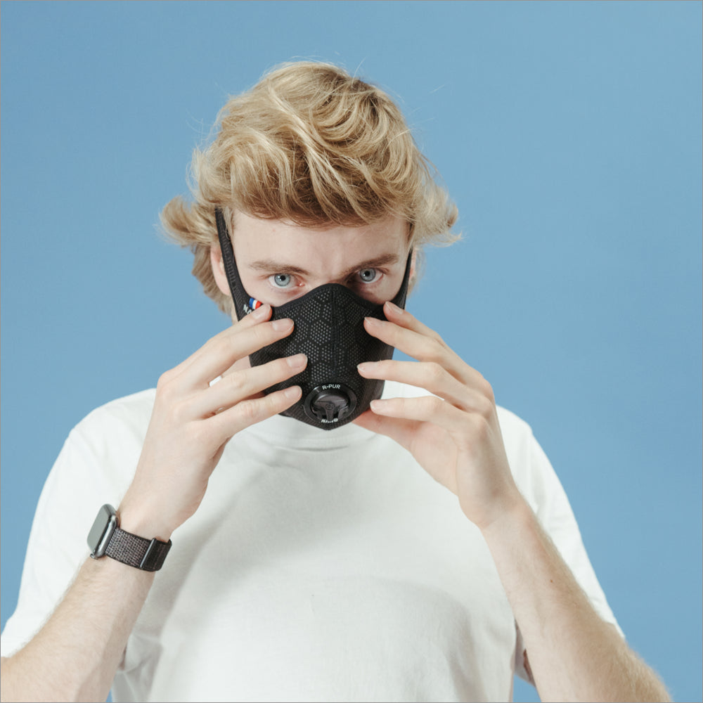 Man in a white T-shirt adjusting a black R-PUR mask against a blue background, putting on his travel mask