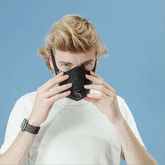 Man in a white T-shirt adjusting a black R-PUR mask against a blue background, putting on his travel mask