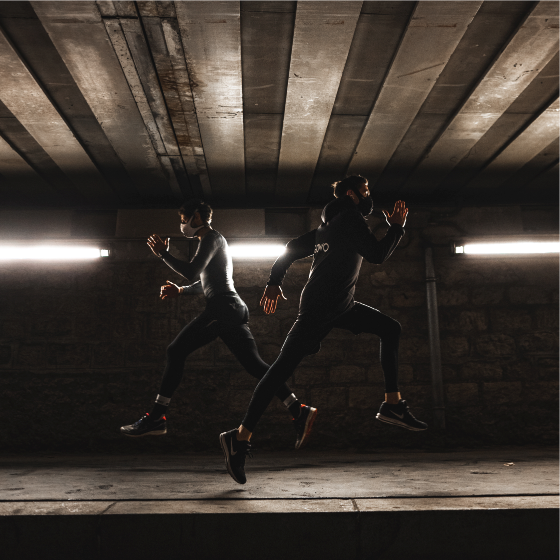 Two athletes sprinting under an overpass, both wearing pollution running masks.