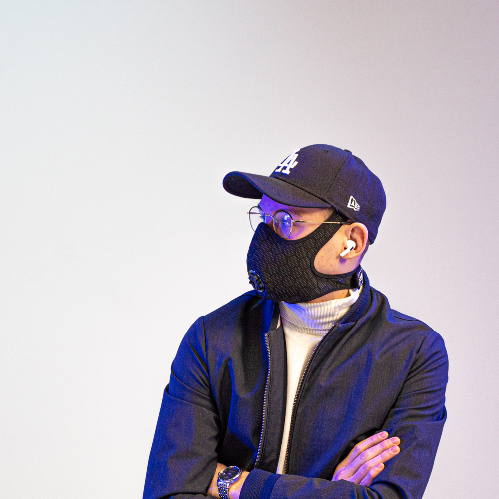 man wearing a travel anti-pollution mask, a cap, and headphones, with his arms crossed and looking to the side