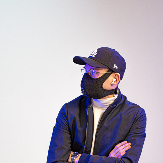 man wearing a travel anti-pollution mask, a cap, and headphones, with his arms crossed and looking to the side
