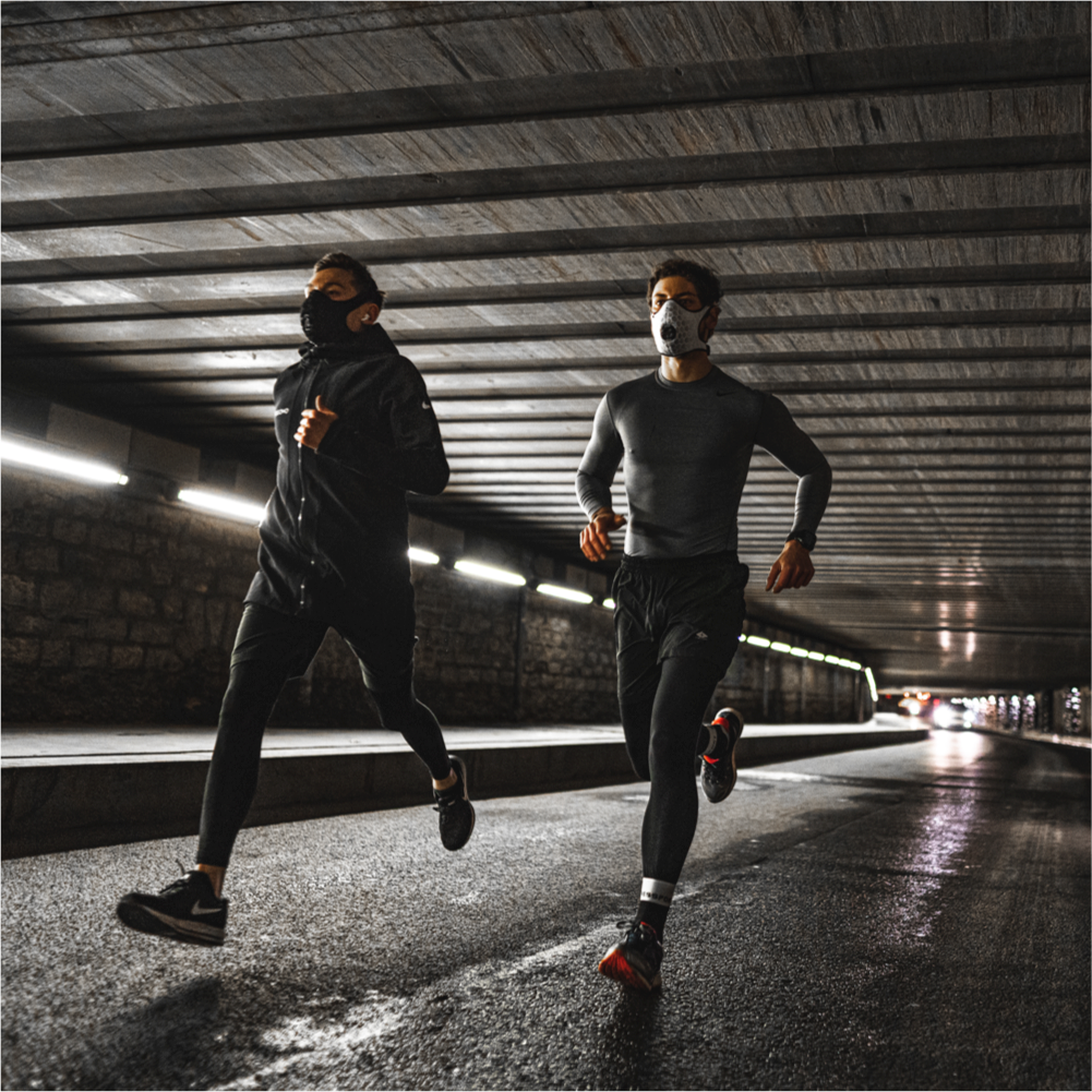 Two runners wearing black and white R-PUR sports masks running under a bridge in the city