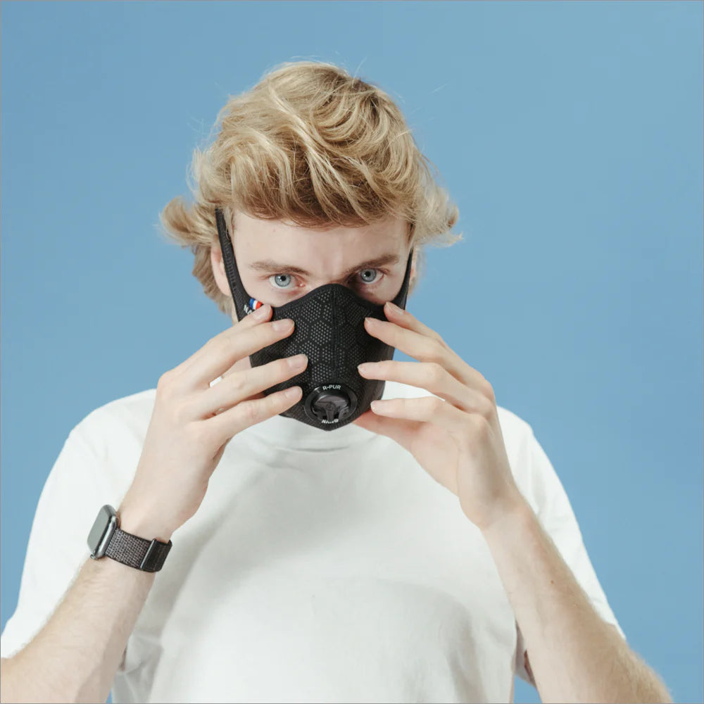 Man in a white T-shirt adjusting a black R-PUR mask against a blue background, putting on his travel mask