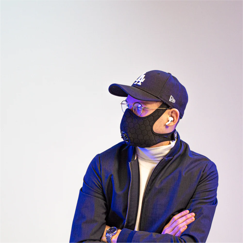 man wearing a travel anti-pollution mask, a cap, and headphones, with his arms crossed and looking to the side